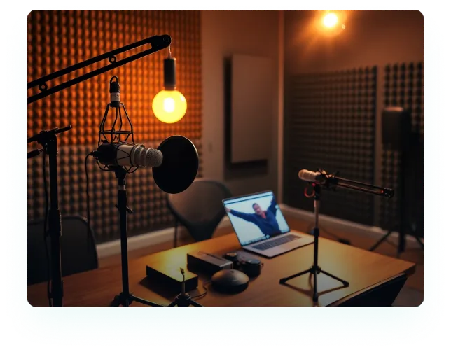 Podcast recording studio with microphones, a laptop showing a person on video call, and warm lighting.